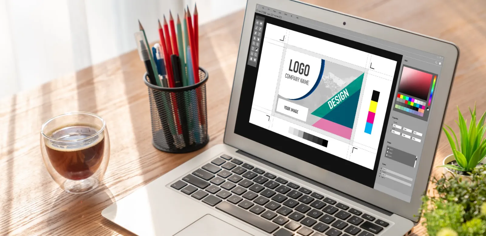 A laptop showing a design layout beside a cup of colored pencils