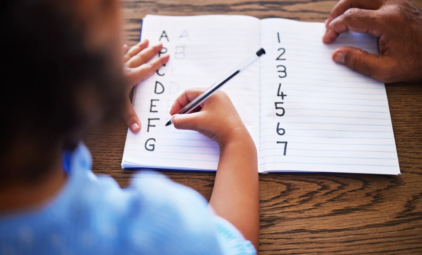 A child writes the alphabet and numbers in a notebook while a teacher's hand rests beside the page