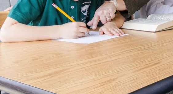 A student writes with a pencil while a teacher points to the page